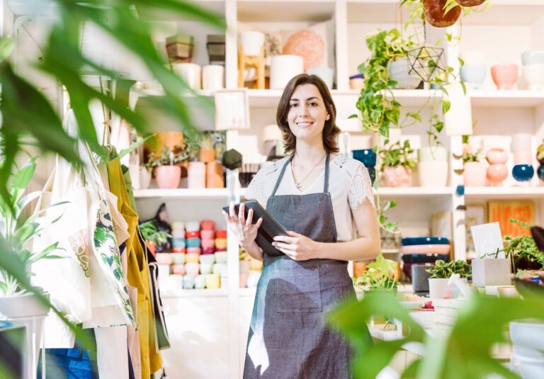 A woman wearing an apron stands in a bright, colorful shop, holding a tablet and smiling—perhaps reviewing her custom website design. Shelves behind her are filled with pots, plants, and ceramics. Green leaves frame the image.