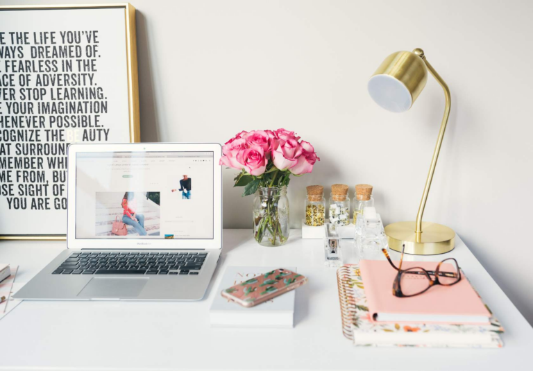 A tidy white desk with a laptop, pink roses in a vase, a gold lamp, notebooks, glasses, and a smartphone—perfect for brainstorming custom website design or logo design services. A framed inspirational quote leans against the wall in the background.