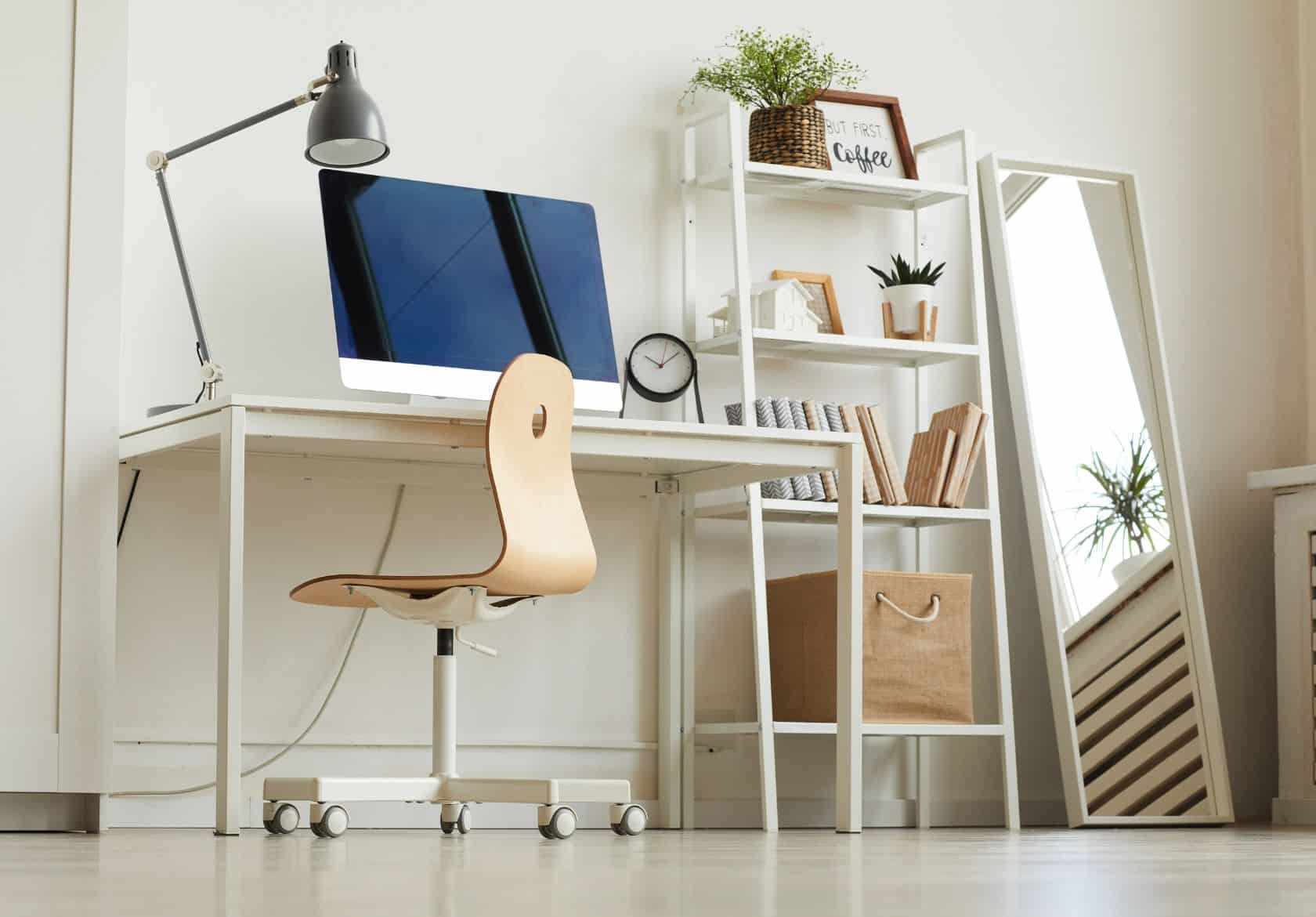 A minimalist home office perfect for a web designer, featuring a wooden chair at a white desk, desktop computer, desk lamp, clock, bookshelf with decor and plants, and a large mirror leaning against the wall.