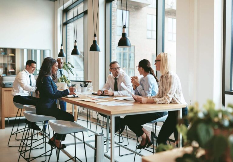 Five people sit around a table in a modern office, engaged in a discussion about custom website design. Papers, notebooks, and laptops are on the table, and large windows let in natural light. The atmosphere appears professional and collaborative.