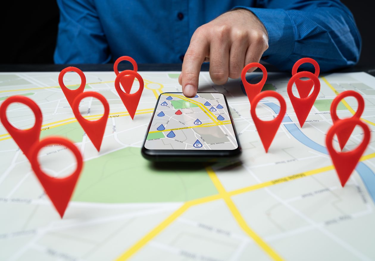 A person uses a smartphone displaying a map with location markers, surrounded by large red map pins on a physical map. Wearing a blue shirt, they point at the phone screen—perfect imagery for showcasing web design services.