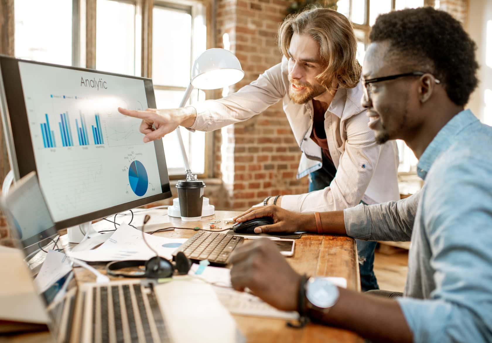 Two men work together at a desk with a computer displaying bar charts and analytics data. One, a skilled web designer from Moonlit Media, points at the screen while the other types, both focused on data analysis in a modern office setting.