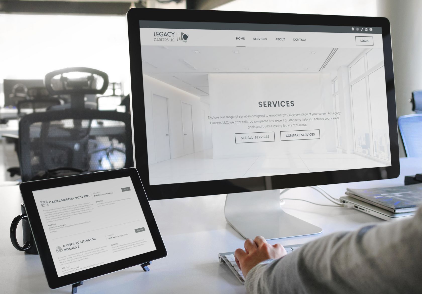 A person uses a desktop computer displaying a website’s Services page, with a tablet nearby showing custom website design options. The workspace features modern office furniture in the background.