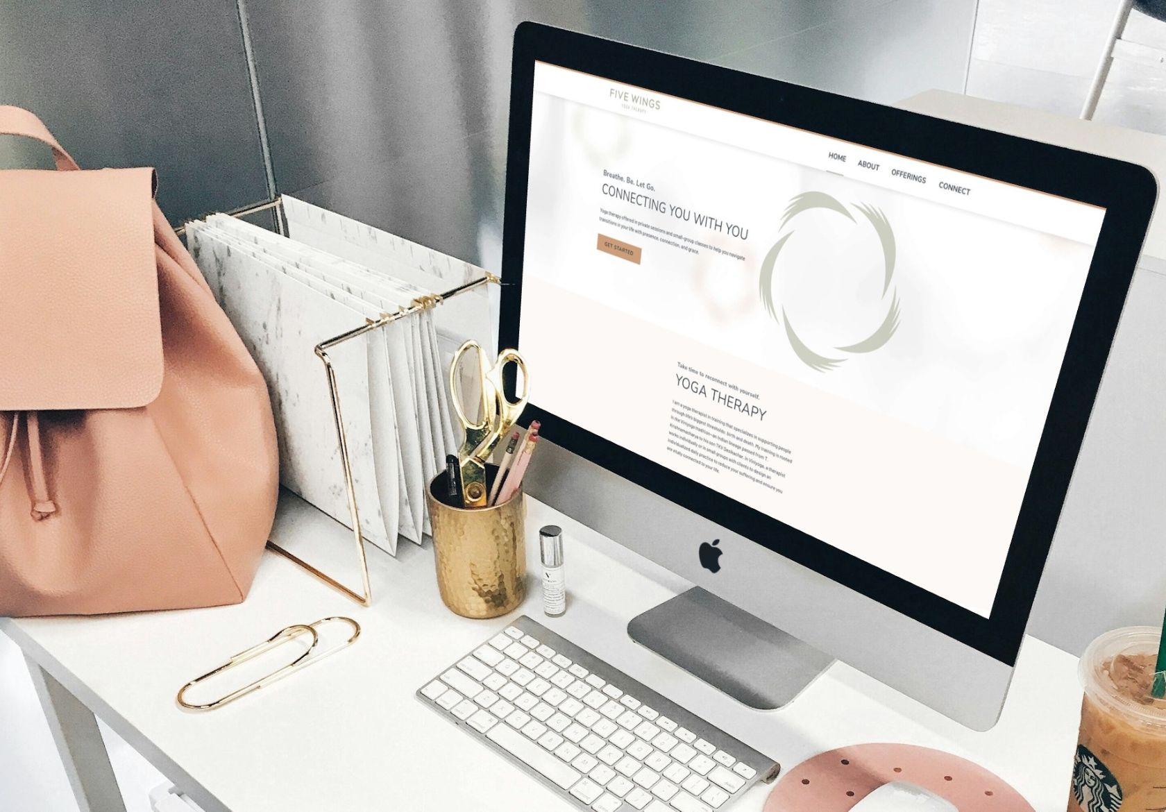 A neat desk with a pink bag, file organizer, gold scissors, and a desktop computer displaying a website about yoga therapy by a web designer. A keyboard, mouse, drink, and office supplies are also on the white desk.
