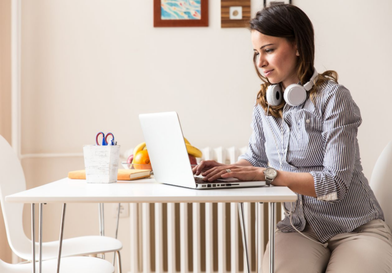 A woman with headphones around her neck works on a laptop at a table, designing a custom logo. Office supplies and fruit are on the table, and framed pictures hang on the wall behind her.