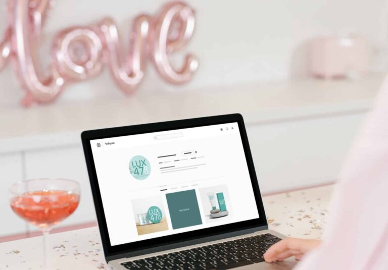 A person uses a laptop displaying a custom website design for LUX 47 skincare products by Moonlit Media. In the background, pink balloon letters spell love, and there’s a glass of pink drink on a confetti-covered table.