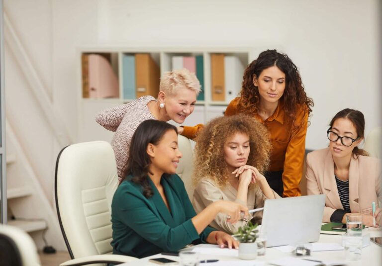 Five women in business attire gather around a desk, collaborating on web design services as they review a laptop screen and documents in a modern office setting.
