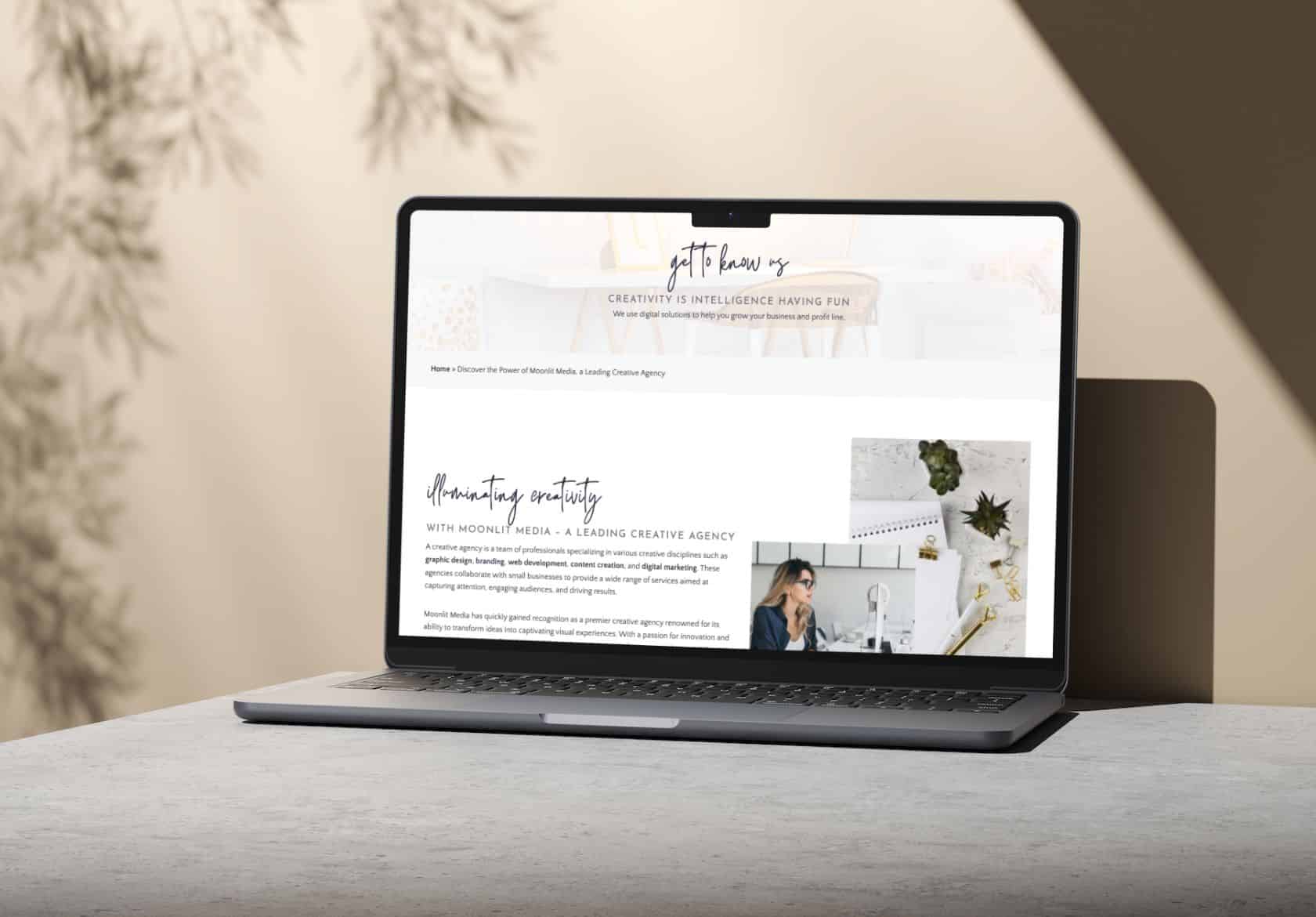 A laptop on a light stone surface displays a website with a creative agency theme, hinting at custom website design. Sunlight casts leaf shadows on the wall, creating a soft, artistic atmosphere.