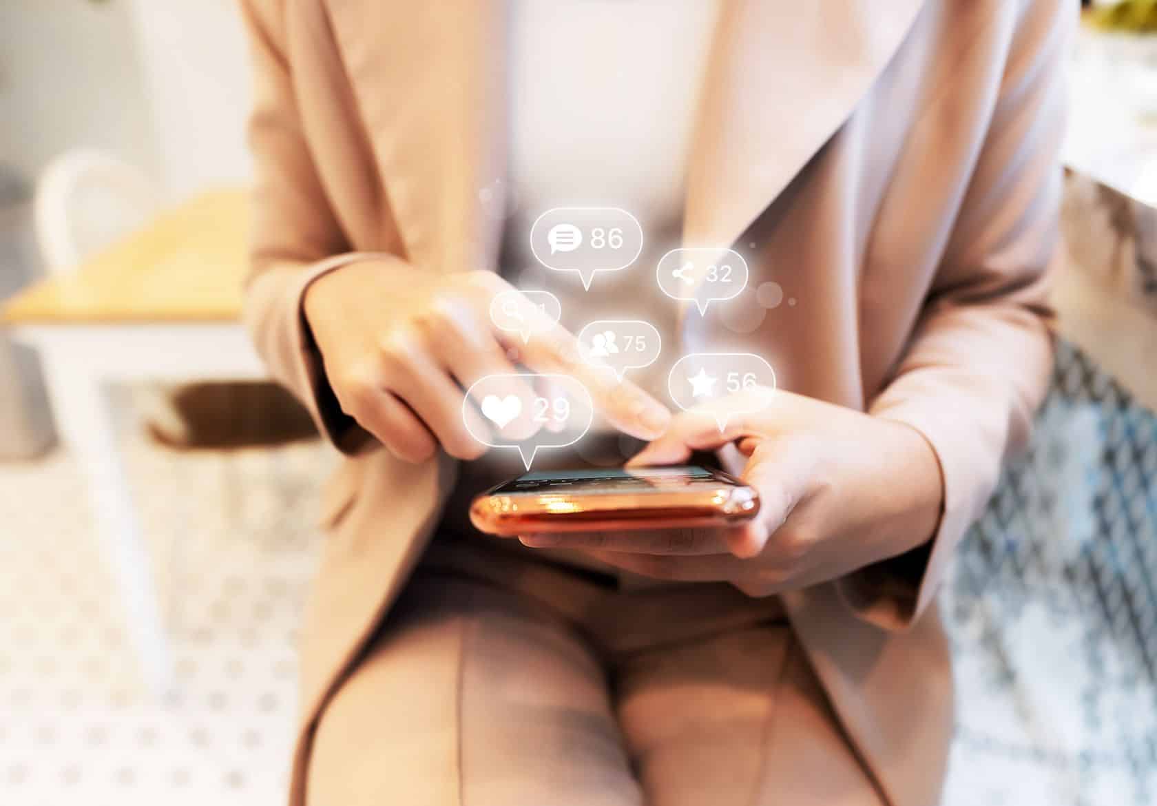 A person in a beige suit uses a smartphone, with social media notification icons like likes, messages, and follows appearing digitally above the device—showcasing Moonlit Media’s expertise in custom website design.