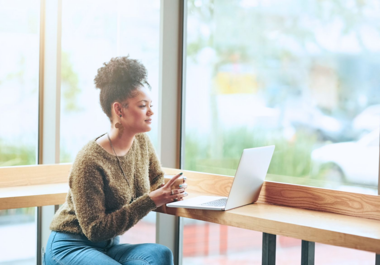 A woman sits by a window at a wooden counter, holding a cup and looking at an open laptop, as natural light streams in and greenery is visible outside—perfectly capturing the workspace of a creative web designer offering web design services.