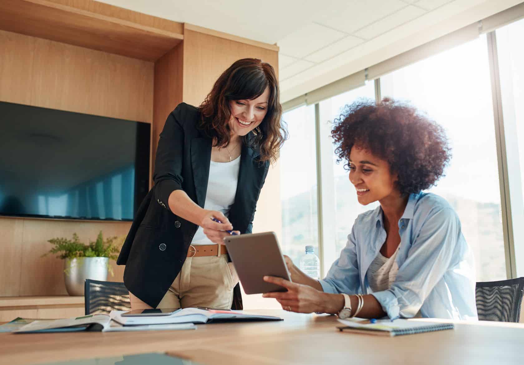 Two women in a modern office, one standing and pointing at a tablet while the other sits, smiling and holding the tablet. They appear to be having a collaborative discussion about logo design services.