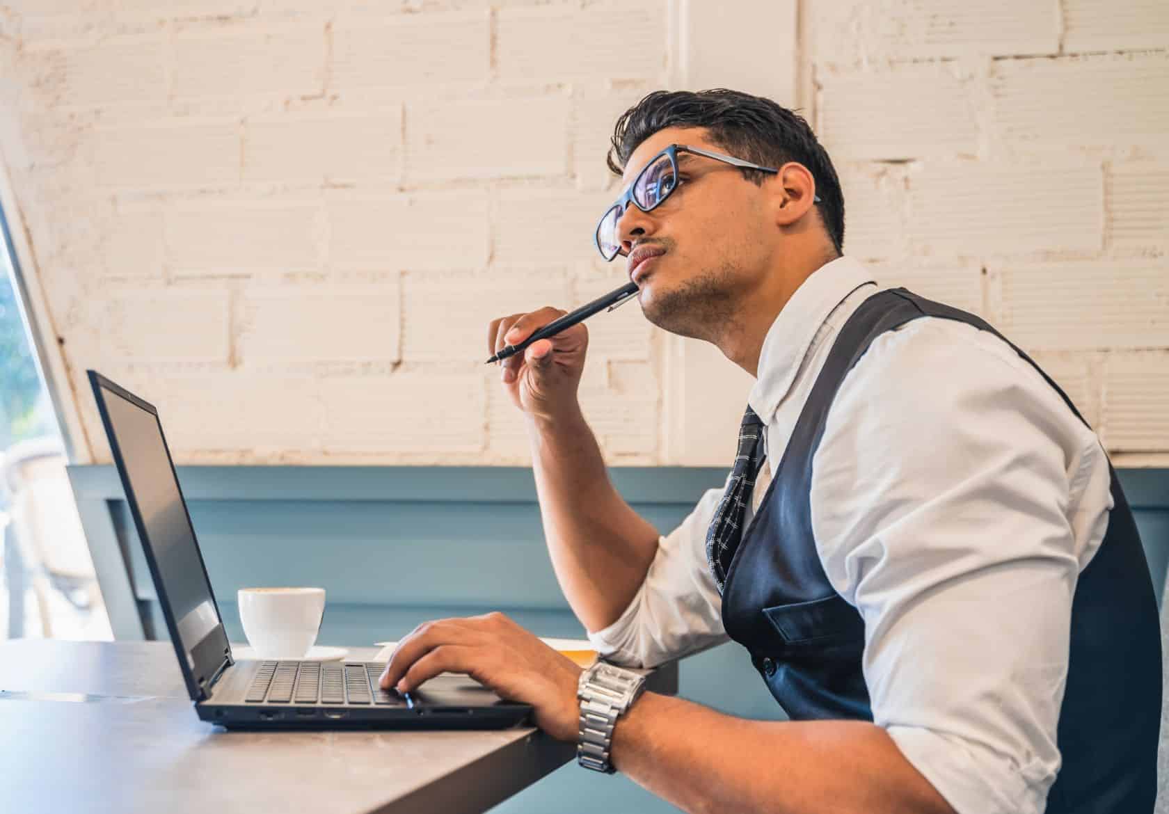 A man in business attire sits at a table with a laptop, looking thoughtful as he considers web design services. A pen is held to his mouth and a white coffee cup rests on the table beside him.