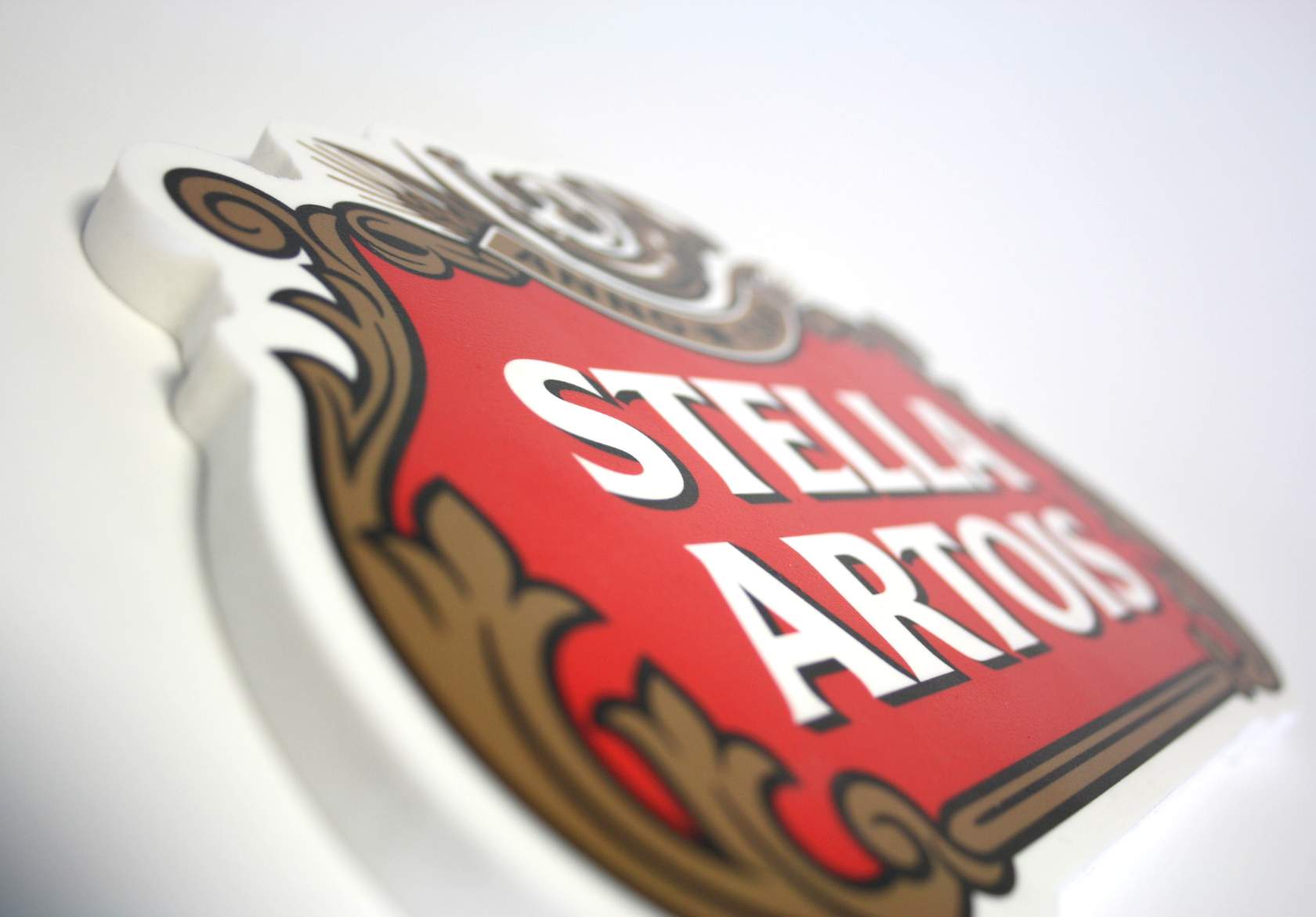 A close-up, angled view of a red and gold Stella Artois logo sign highlights custom logo design on a white background, with the text partially in focus and decorative scrollwork visible.