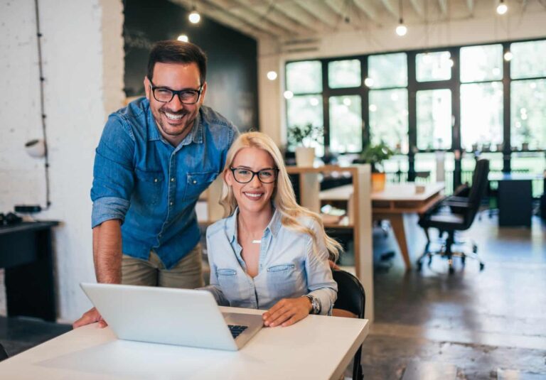 A man and a woman, both wearing glasses and denim shirts, smile at the camera while collaborating on custom logo design in a modern, bright office space with large windows and exposed ceiling beams.