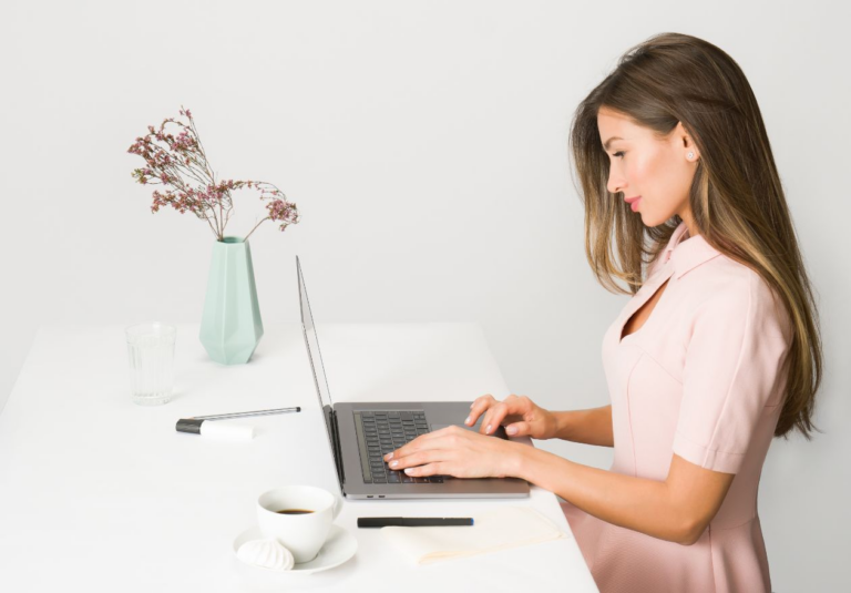 A woman in a light pink dress sits at a white desk, working on custom logo design. On the desk are a cup of coffee, a saucer with meringues, a pen, glasses, and a mint-green vase with dried flowers.