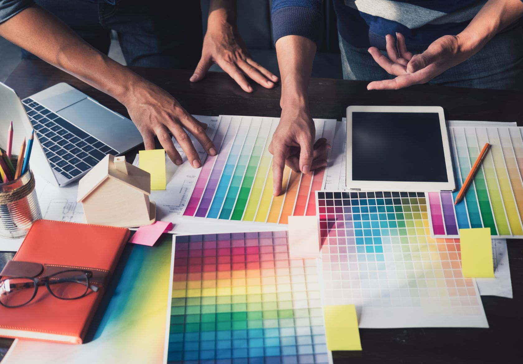 Two people discuss colorful swatches and design ideas at a table covered with color charts, a digital tablet, a laptop, notebooks, pencils, and sticky notes—perfect for planning custom website design with Moonlit Media.