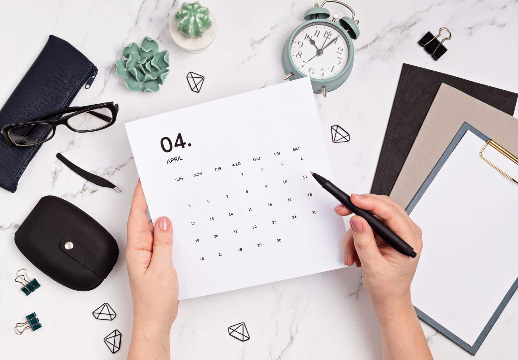 A person holds a calendar for April while pointing at it with a pen. Nearby are eyeglasses, a clock, stationery, and a potted plant on a marble desk—an inspiring setup for planning custom website design with Moonlit Media.