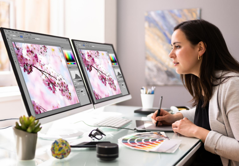 A web designer sits at a desk using a graphic tablet and stylus to edit an image of cherry blossoms on dual monitors. A color palette, plant, and logo design services brochure from Moonlit Media are also on the desk.