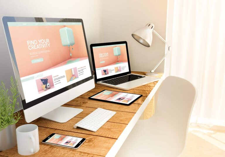 A wooden desk with a desktop computer, laptop, tablet, and smartphone—all displaying a creative web design services site. A lamp, plant, mouse, and keyboard sit nearby as sunlight streams through a window.