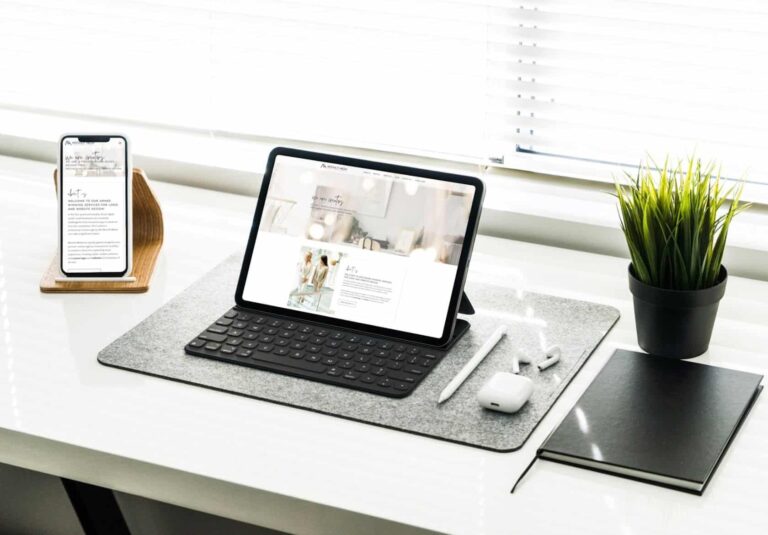 A tablet with a keyboard, a smartphone on a wooden stand, a potted plant, AirPods, and a notebook are neatly arranged on a white desk near a window—an inspiring setup for custom website design projects by Moonlit Media.