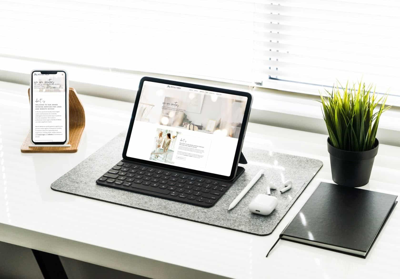 A tablet with a keyboard, a smartphone on a wooden stand, a potted plant, AirPods, and a notebook are neatly arranged on a white desk near a window—an inspiring setup for custom website design projects by Moonlit Media.