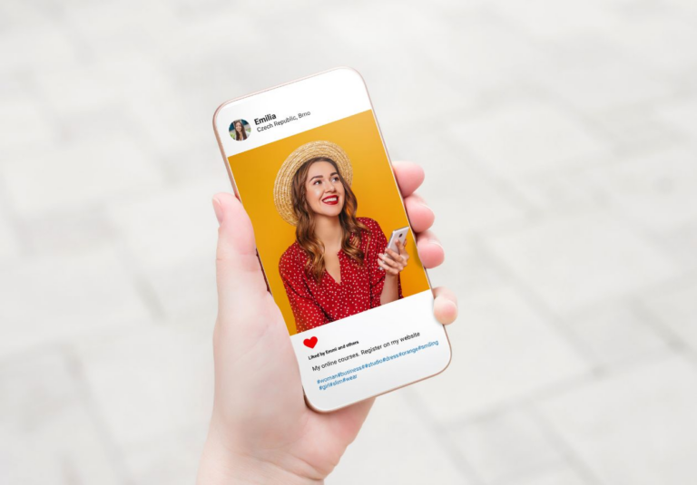 A hand holds a smartphone displaying a social media post from Moonlit Media, showing a smiling woman in a straw hat and red polka-dot dress against a yellow background. The post features a heart emoji and highlights their custom logo design service.