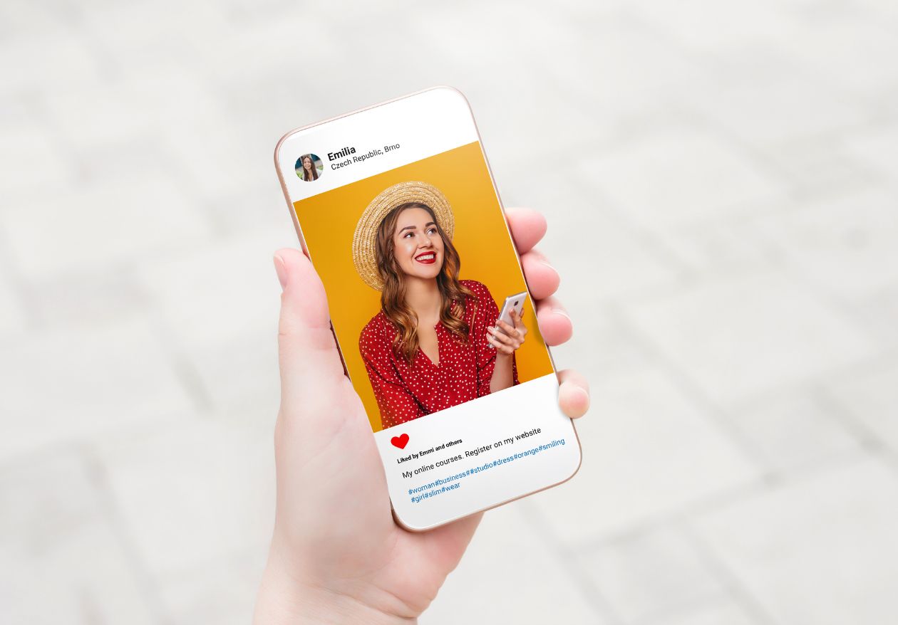 A hand holds a smartphone displaying a social media post from Moonlit Media, showing a smiling woman in a straw hat and red polka-dot dress against a yellow background. The post features a heart emoji and highlights their custom logo design service.