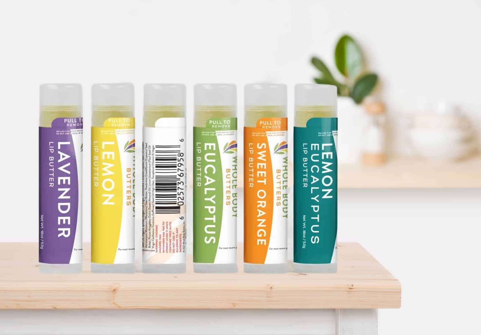 Six lip butter tubes in assorted colors and flavors—lavender, lemon, eucalyptus, lemon eucalyptus, and sweet orange—stand upright on a light wooden surface, paired perfectly with a moonlit media custom logo design featured in the blurred background.