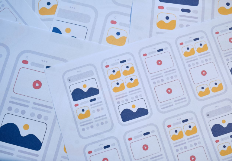 Sheets of paper display various mobile app interface wireframes, featuring image and video placeholders in blue, yellow, and red on white backgrounds, illustrating different layout designs often used by web design services.