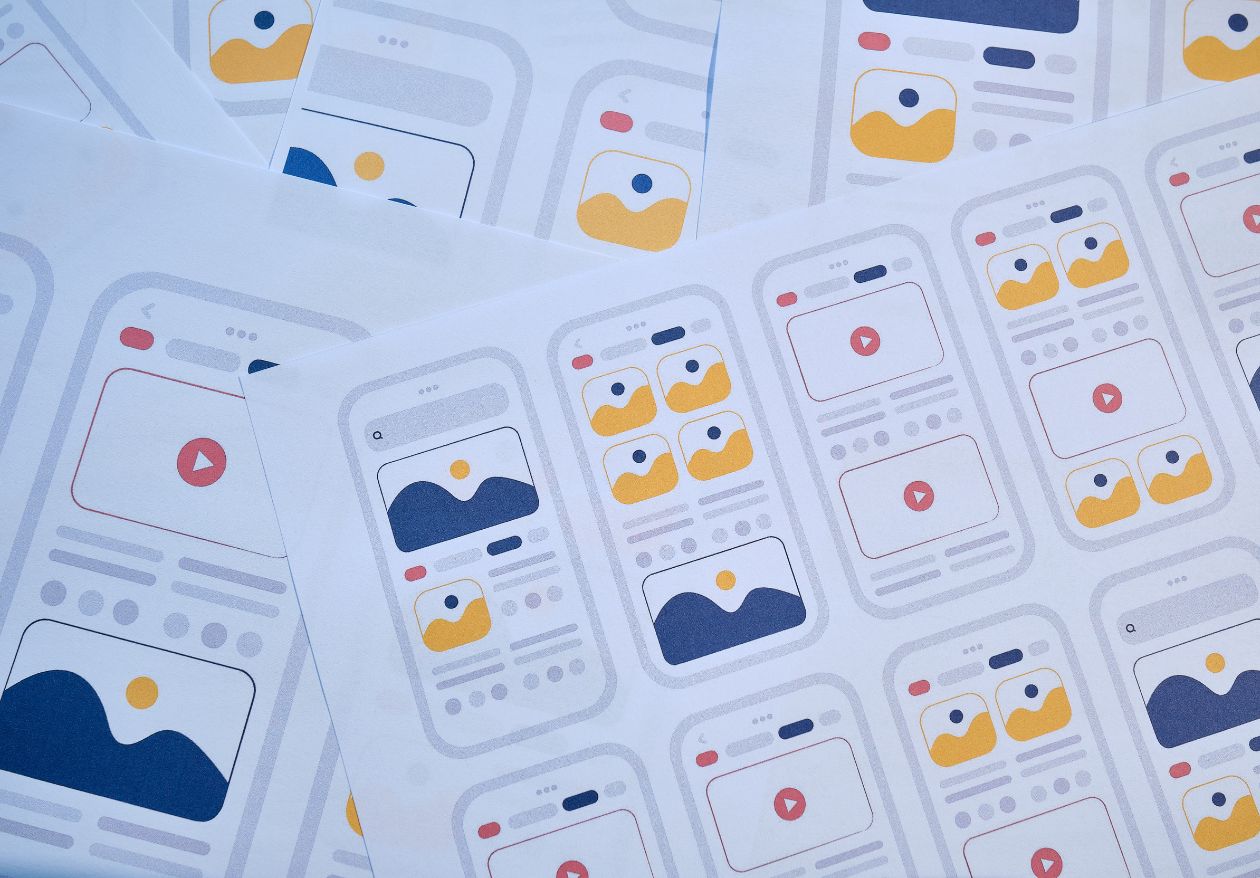 Sheets of paper display various mobile app interface wireframes, featuring image and video placeholders in blue, yellow, and red on white backgrounds, illustrating different layout designs often used by web design services.