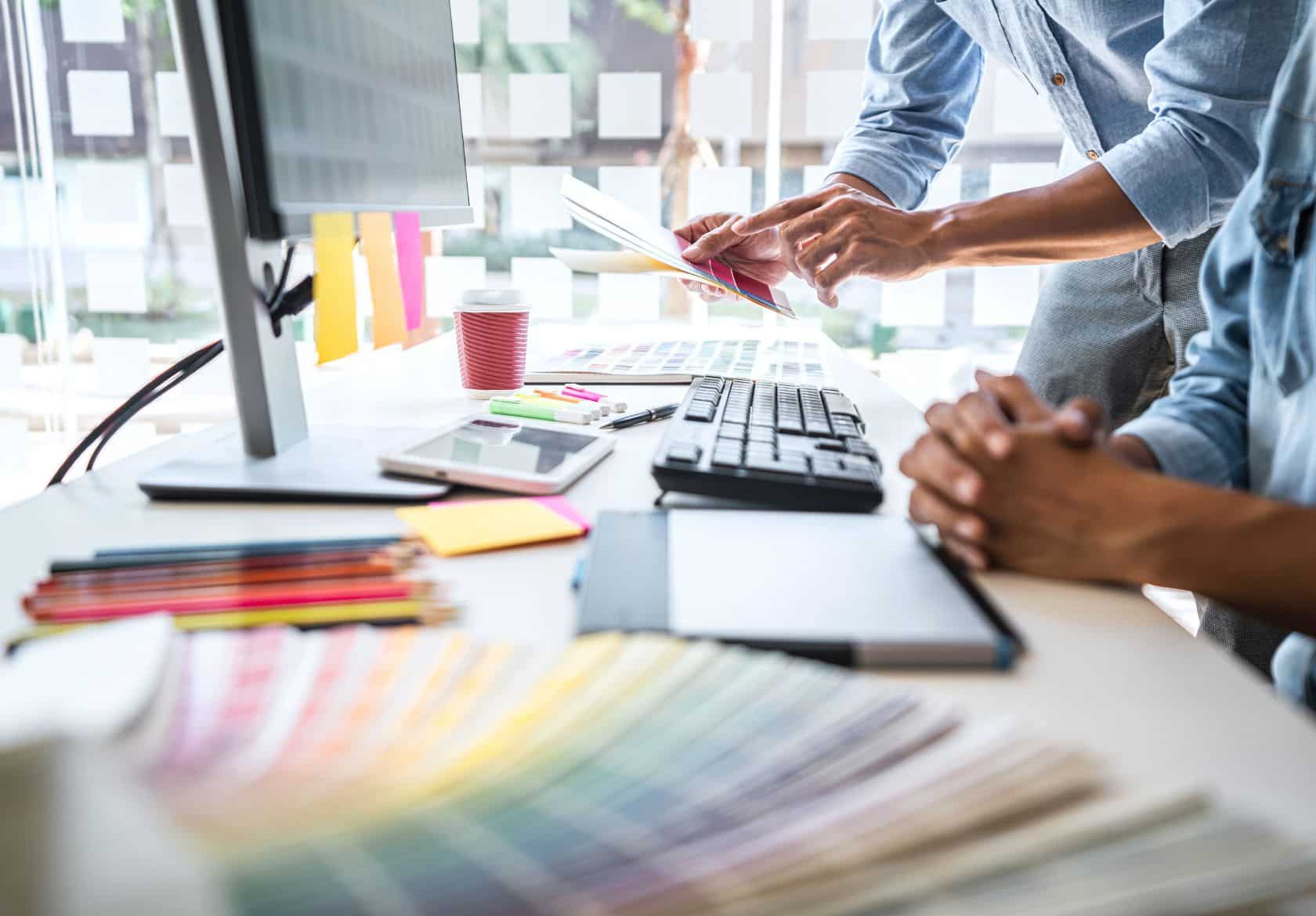 Two people work at a desk with a computer, color swatches, and notepads, discussing papers—showcasing Moonlit Media’s collaborative approach to custom logo design and web design services.