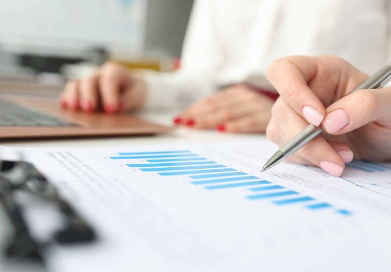 Close-up of a hand holding a pen, pointing at a blue bar graph on a printed document—perfect for a web designer planning custom website design. In the background, another person types on a laptop. Both have manicured nails.