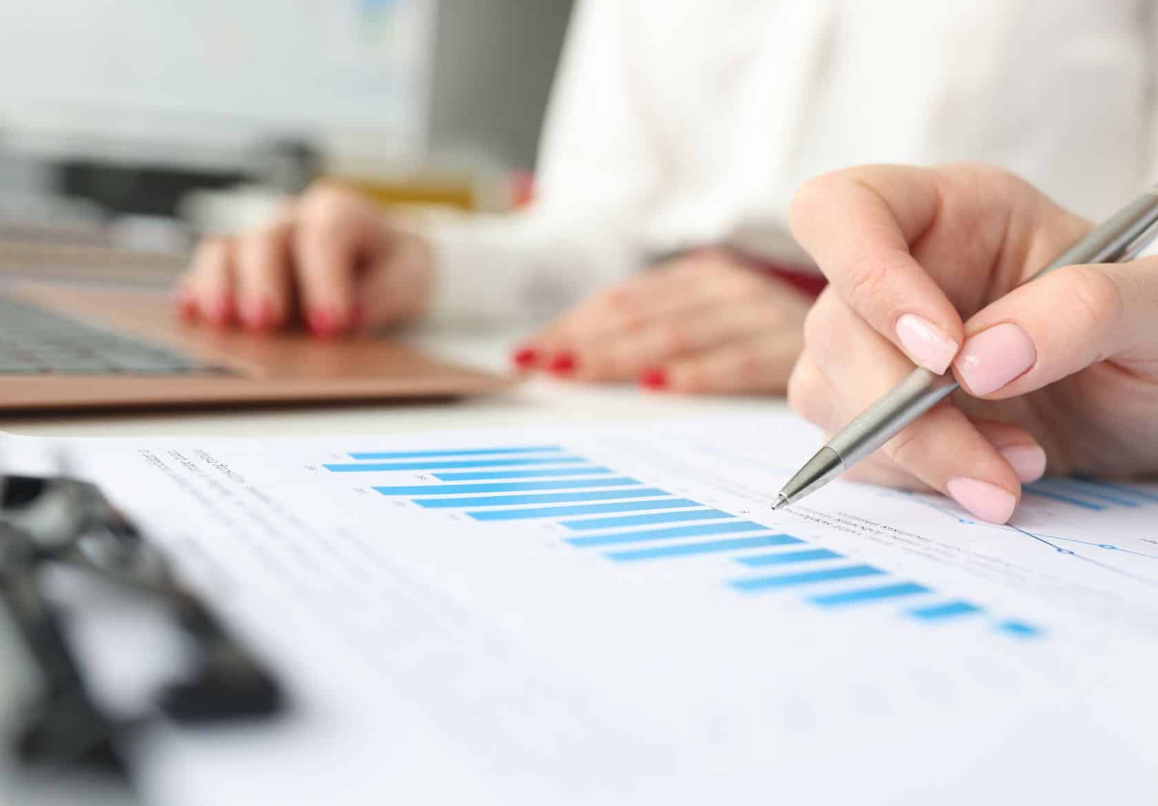 Close-up of a hand holding a pen, pointing at a blue bar graph on a printed document—perfect for a web designer planning custom website design. In the background, another person types on a laptop. Both have manicured nails.