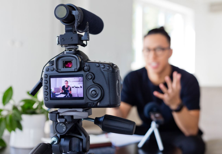 A camera on a tripod records a person indoors, speaking and gesturing while discussing custom website design. The person appears on the camera’s screen, with a microphone and a plant visible in the background.
