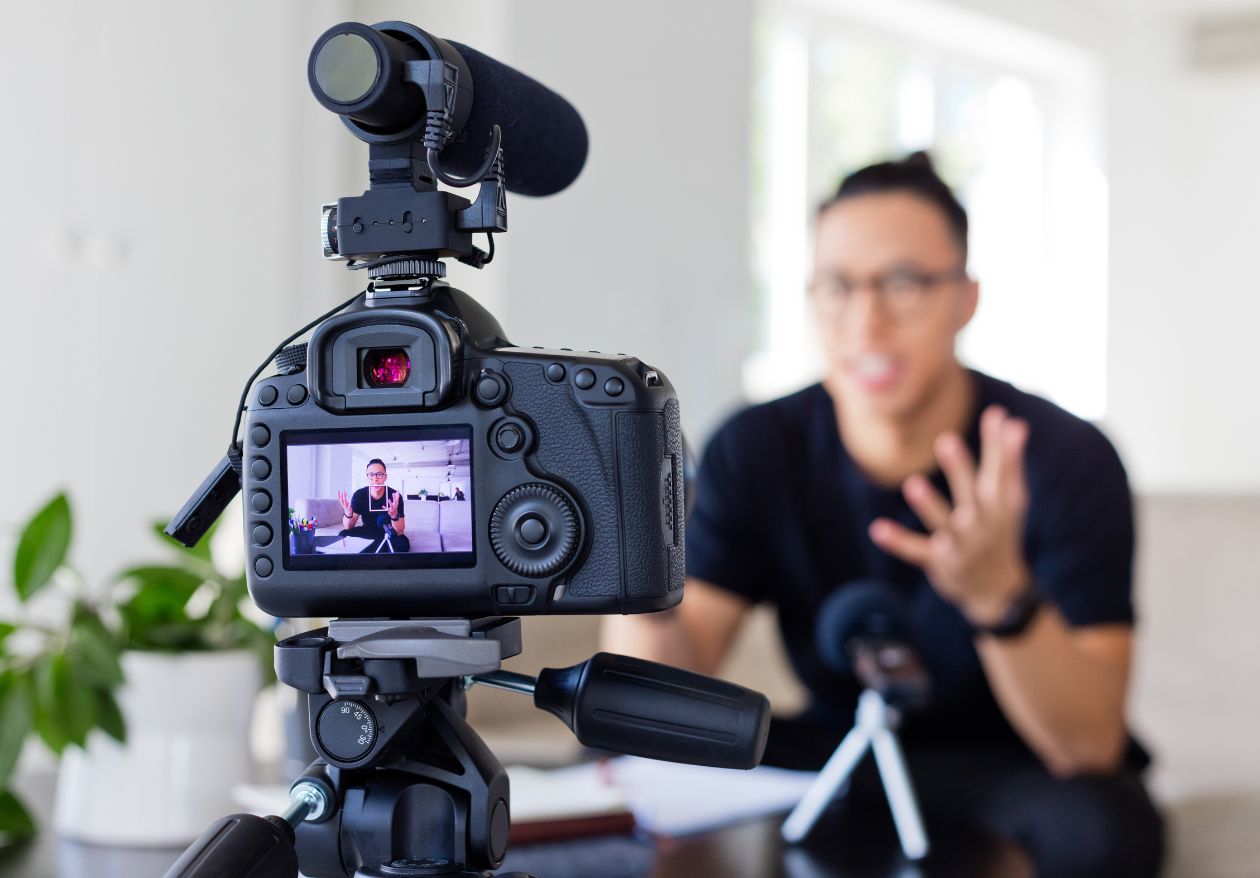 A camera on a tripod records a person indoors, speaking and gesturing while discussing custom website design. The person appears on the camera’s screen, with a microphone and a plant visible in the background.