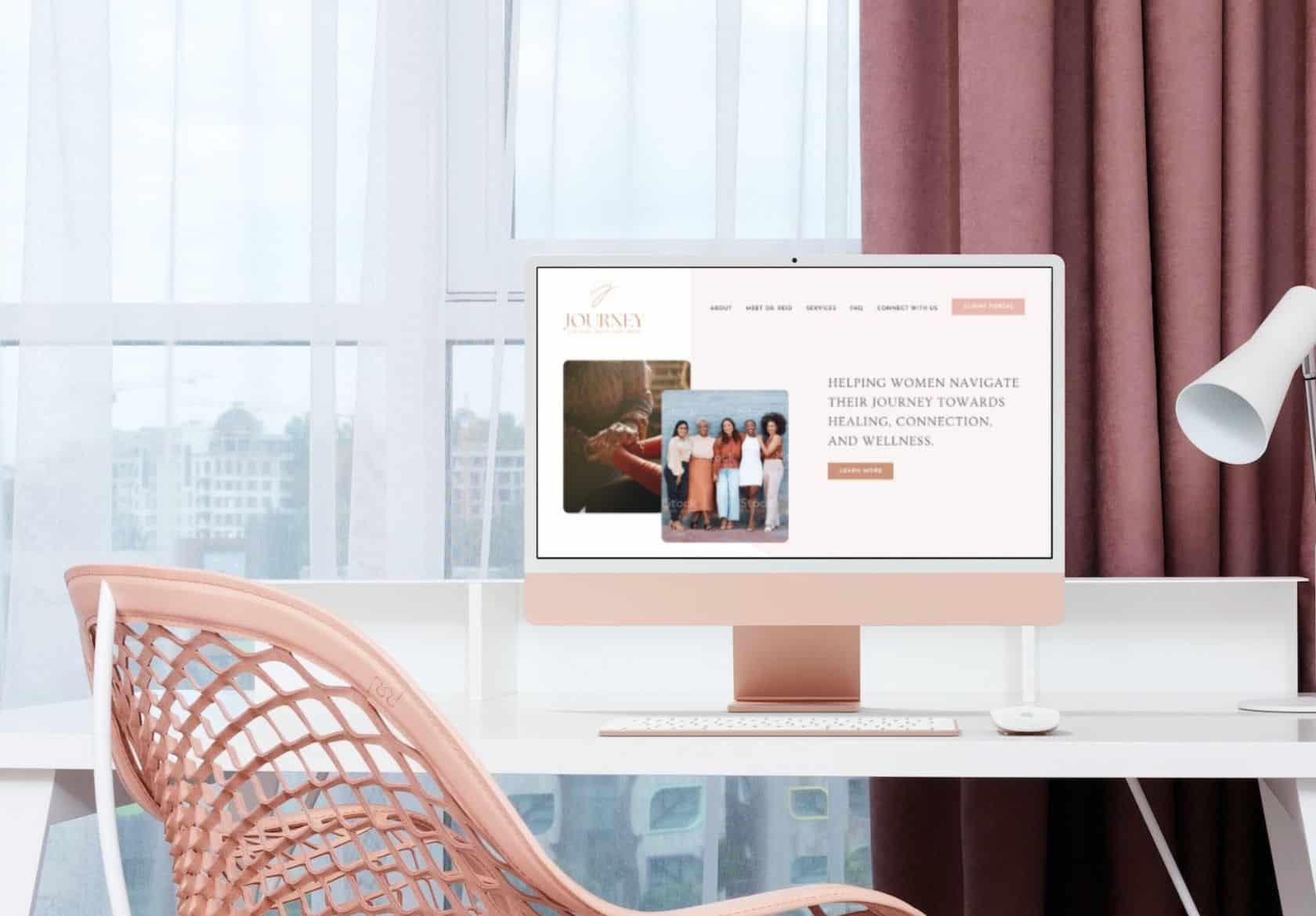 A modern home office with a blush pink chair and a white desk featuring a desktop computer displaying a web designer’s site about helping women with healing, connection, and wellness. City buildings are visible through the window.