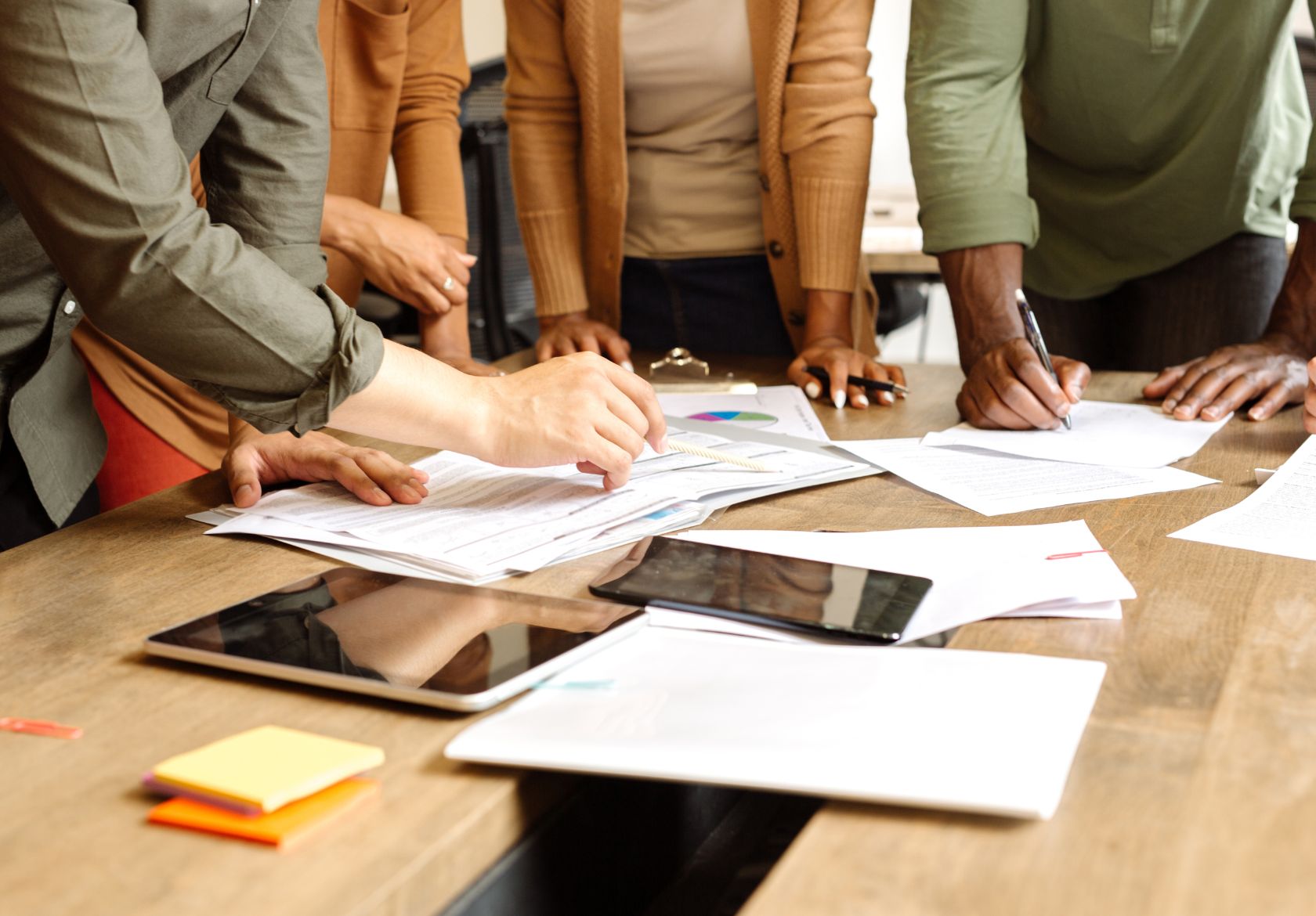 A group of people stands around a wooden table, reviewing documents and charts. Papers, a tablet, and sticky notes are spread out as they discuss custom website design strategies for Moonlit Media’s web design services.