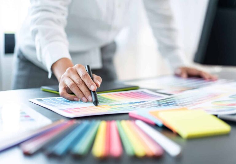 A web designer in a white shirt uses a stylus to point at a color swatch chart on a desk, surrounded by colorful markers, notepads, and design materials—brainstorming ideas for custom website design and logo design services.
