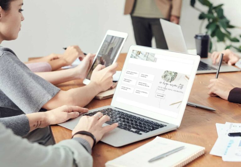 People sitting at a table in a meeting, using laptops and tablets. A web designer stands in the background while others work and take notes. A presentation on custom website design is visible on one of the laptop screens.