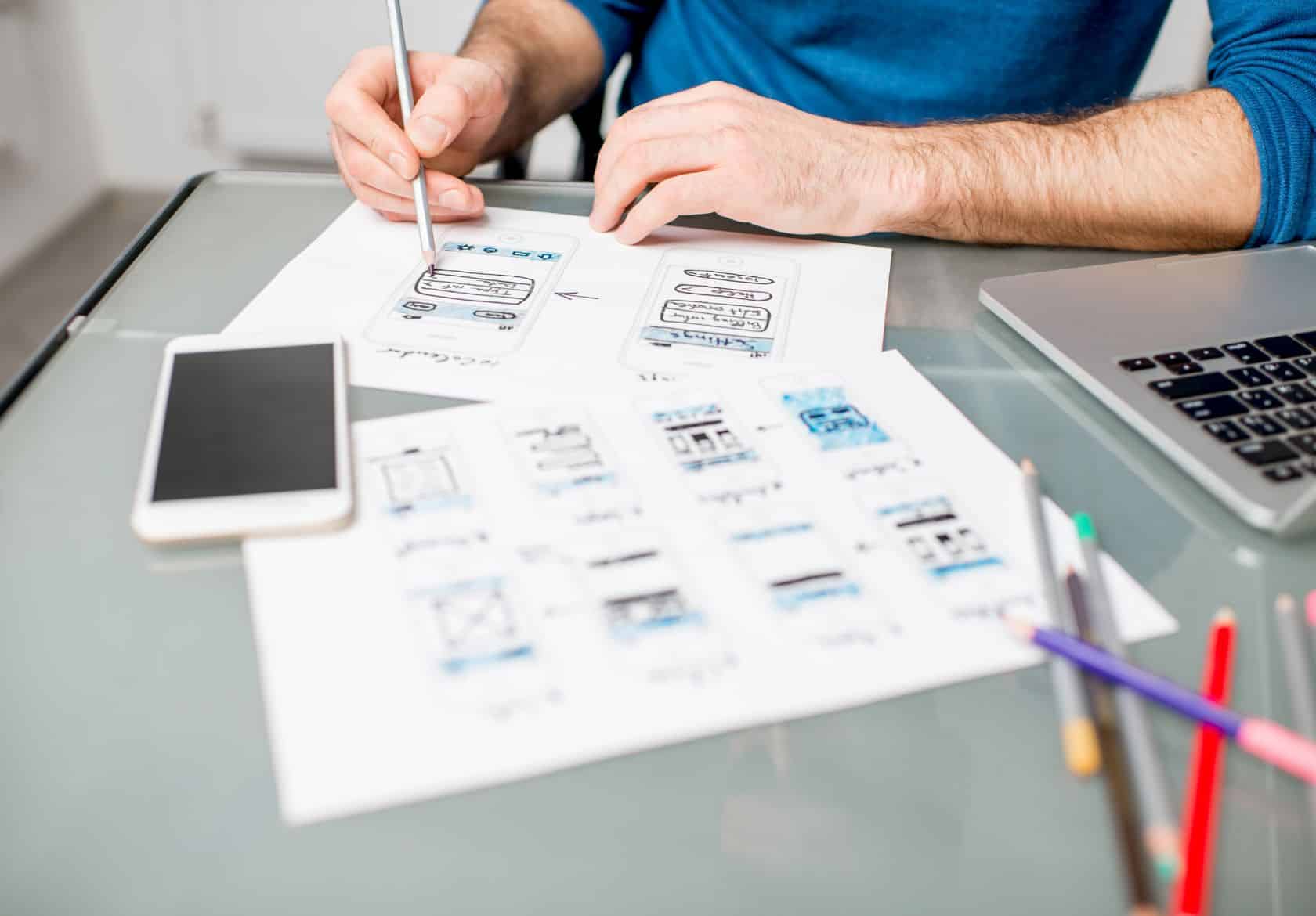 A person sketches mobile app wireframes on paper at a desk, surrounded by a smartphone, colored pencils, and a laptop—perfectly capturing the creative process behind custom website design and logo design services at Moonlit Media.