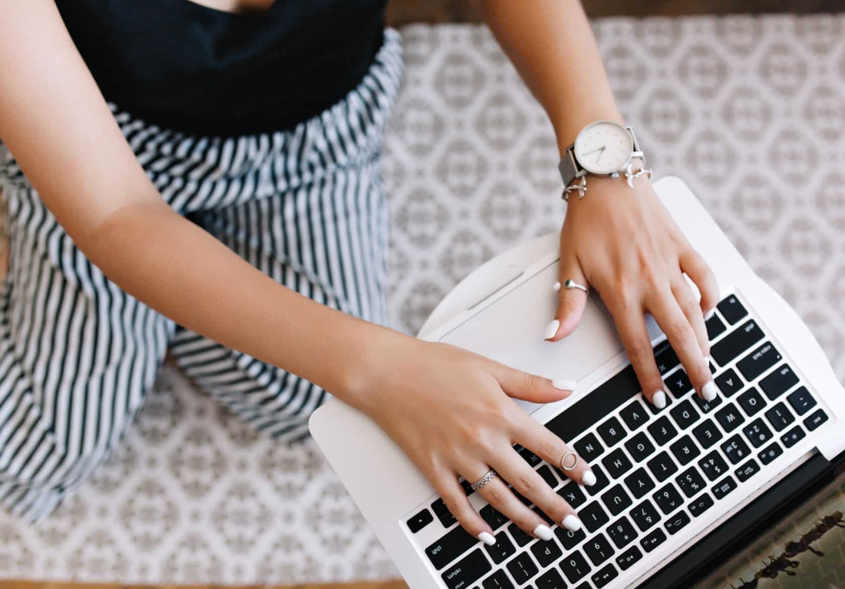 A web designer wearing striped pants and a watch types on a laptop, sitting on a patterned floor. Their nails are painted white, and the image is viewed from above.