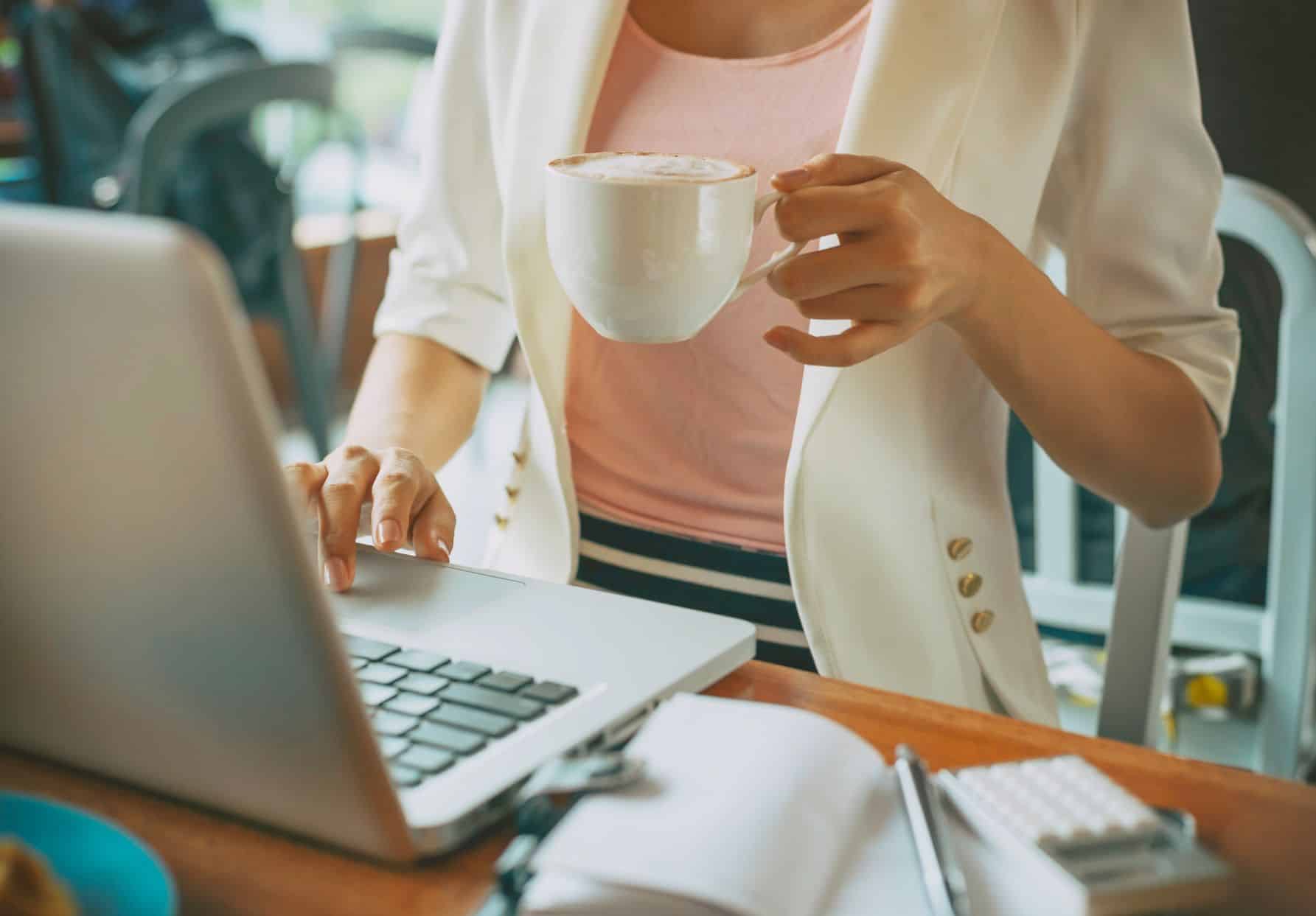 A person in a white blazer holds a cup of coffee while working on custom website design at a wooden table, with a notebook, pen, and calculator nearby.