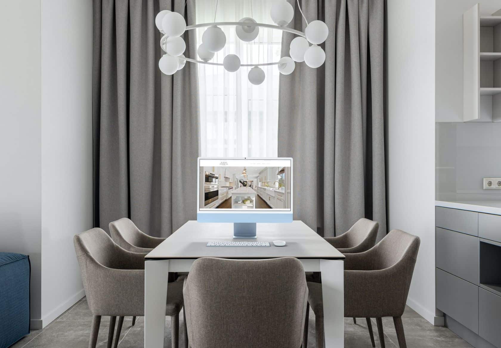 A modern dining room with a white table, six beige chairs, a large window with gray curtains, and a contemporary chandelier. A computer monitor on the table displays a custom website design.