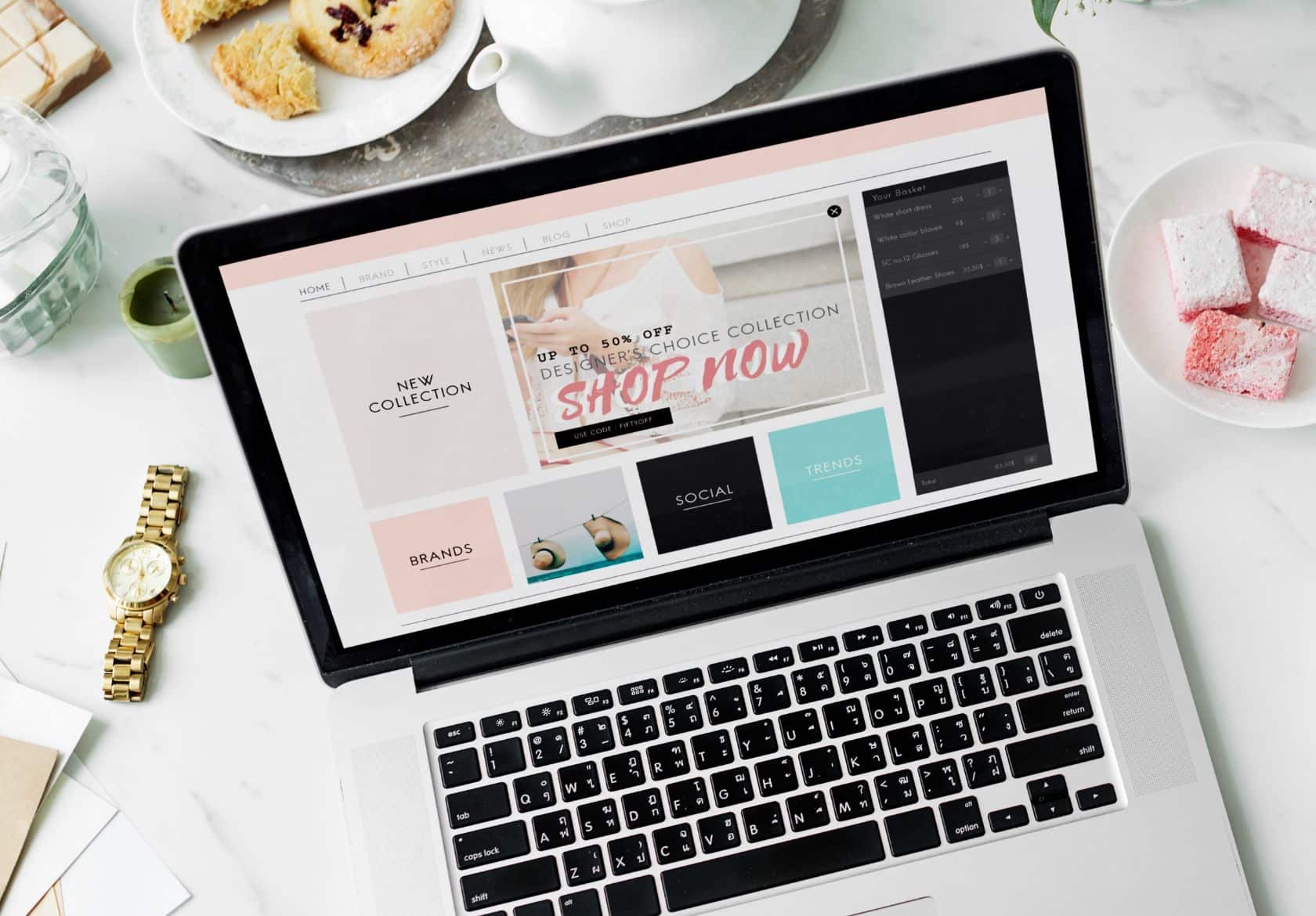 A laptop on a table displays an online shop with a Shop Now banner, highlighting expert web design services. Surrounding the laptop are a gold watch, a glass of water, pastries, and pink marshmallows on a white plate.