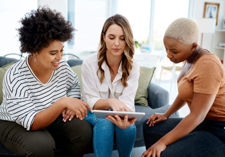 Three women sit together on a couch, looking at a tablet device. They appear engaged and focused, discussing or viewing custom logo design options in a bright, modern living room.