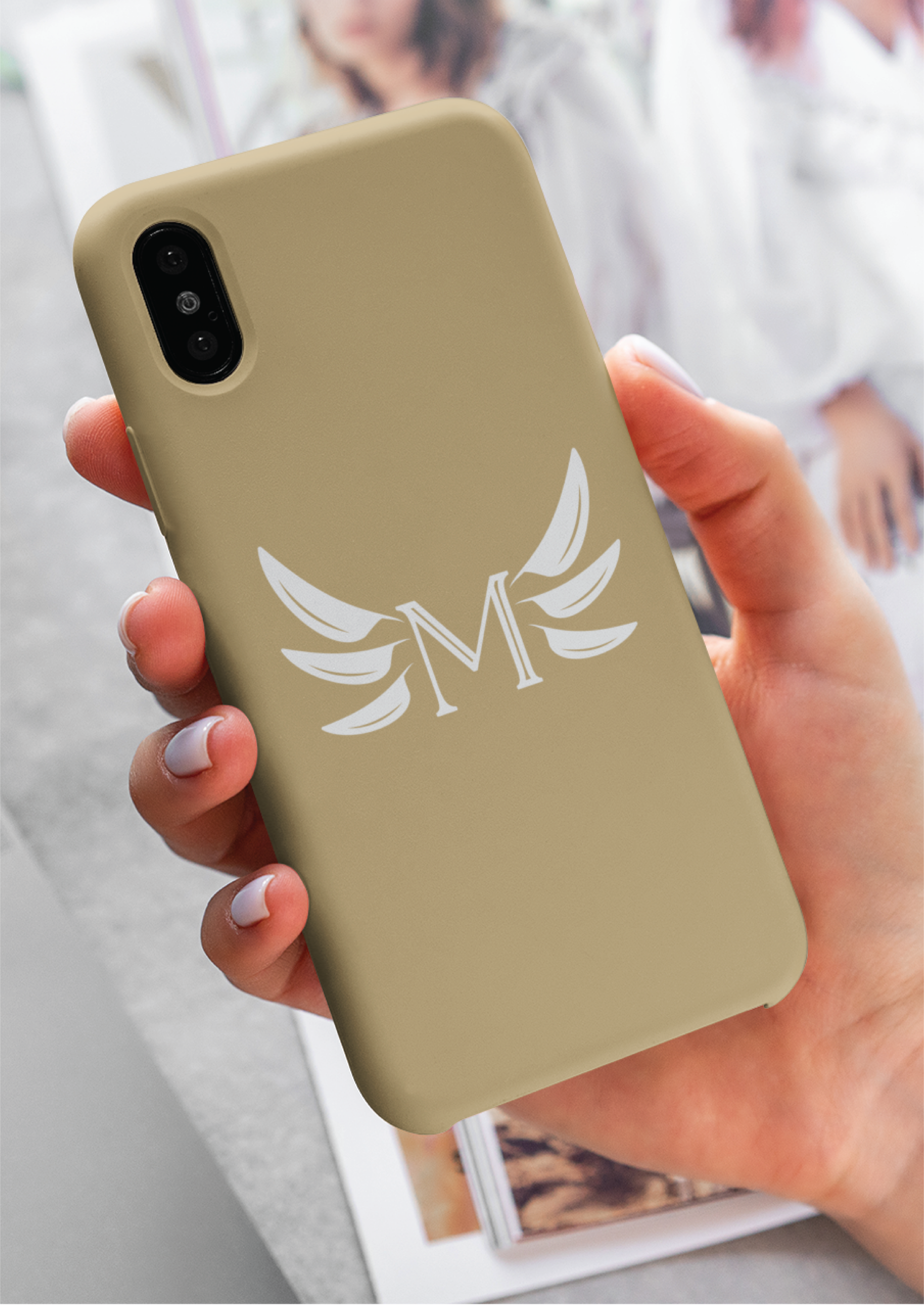 A hand holds a beige phone case featuring a white letter M with stylized wing designs, reflecting sleek branding. In the background, a blurred magazine with images adds depth to the scene.
