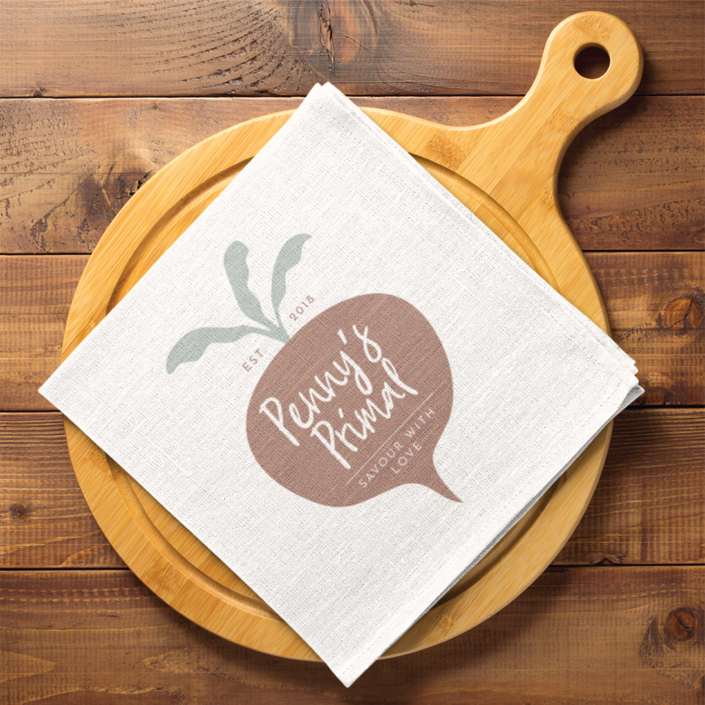 A white cloth napkin with a turnip illustration and the text “Penny’s Primal, Savoured with Love” showcases charming branding for the home cook, resting on a round wooden cutting board atop a rustic wooden table.