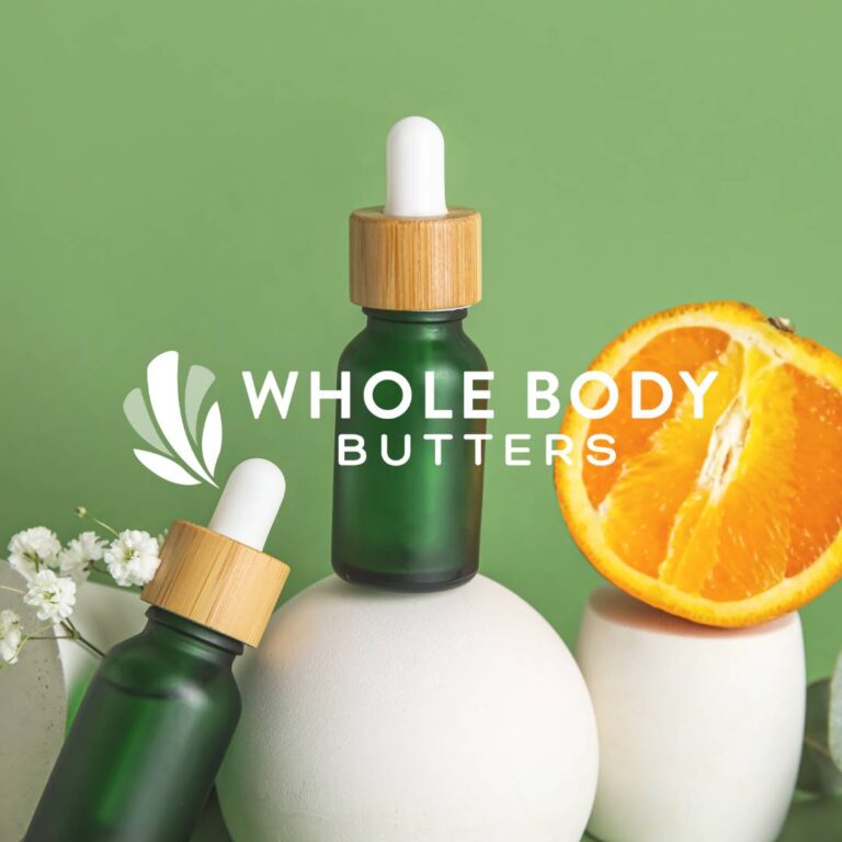 Two green glass dropper bottles with wooden caps are displayed among white round props, a halved orange, and white flowers on a green background. The text reads “Whole Body Butters” by Moonlit Media, specialists in custom website design.