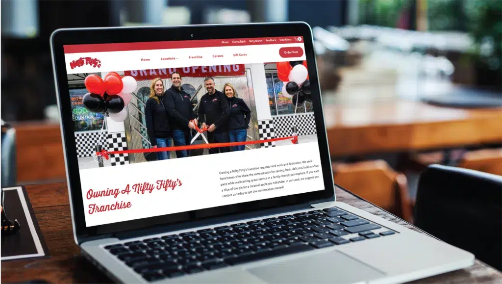 website-design-for-franchise-restaurant-business-1024x579.png | Moonlit Media LLC A laptop on a wooden table displays a website redesign featuring four people at a ribbon-cutting event with balloons and a Grand Opening banner, promoting a Nifty Fiftys franchise in New Hampshire.