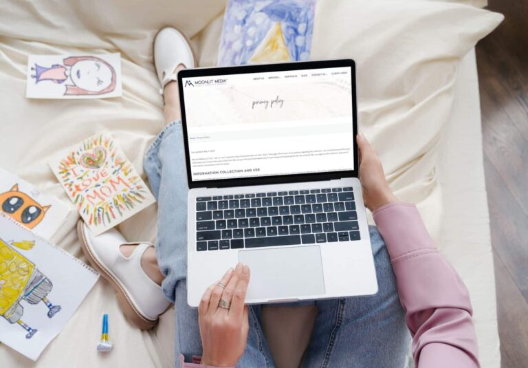 A person sits cross-legged on a couch, holding a laptop surrounded by colorful drawings and art supplies. The screen displays the Mount Meche site—an example of custom website design by Moonlit Media.